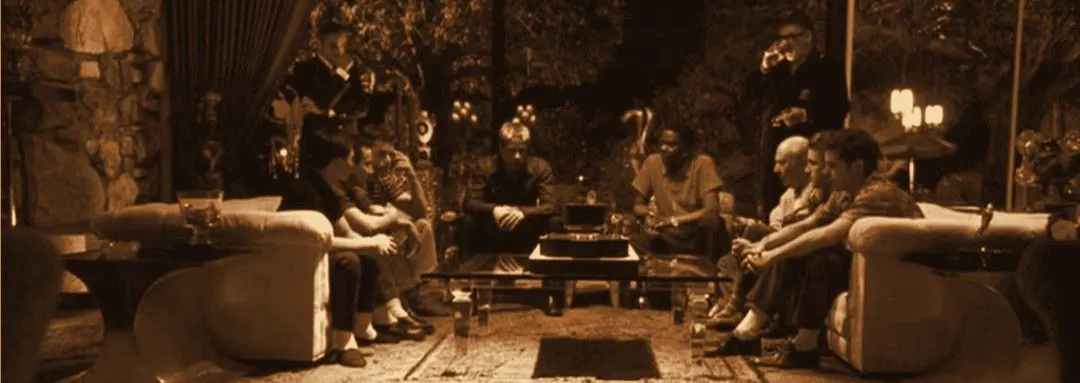 The crew sitting around a living room at night, tungsten tone, wide frame